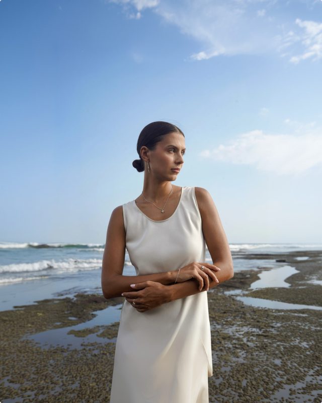 Frau im hellen Kleid mit LIOR-Schmuck an der Küste, blauer Himmel